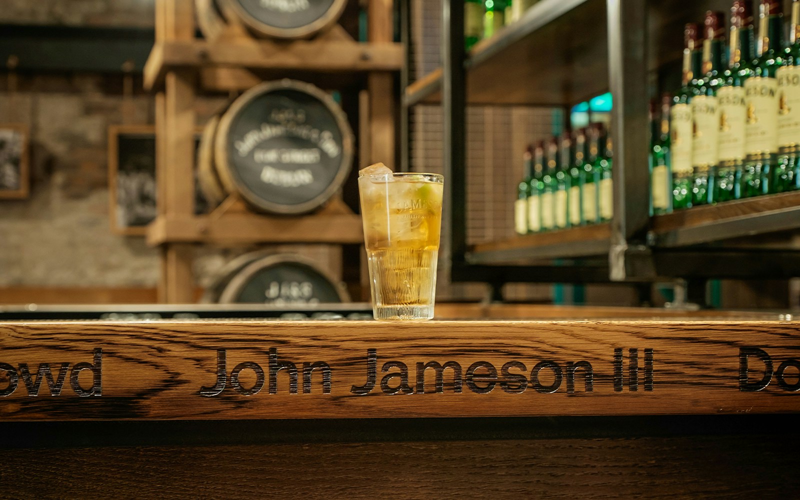 Glass of whiskey on bar at Jameson Distillery with barrels and bottles in background.