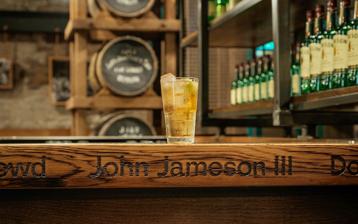 Glass of whiskey on bar at Jameson Distillery with barrels and bottles in background.
