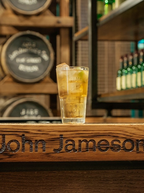 Glass of whiskey on bar at Jameson Distillery with barrels and bottles in background.