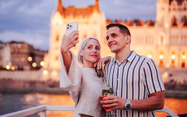Couple taking selfie on Budapest Danube River cruise with Parliament building in background.