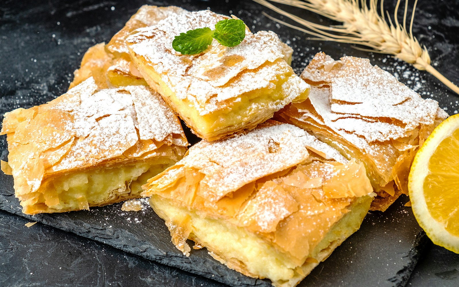 Greek bougatsa pastry with powdered sugar and lemon slice on slate.