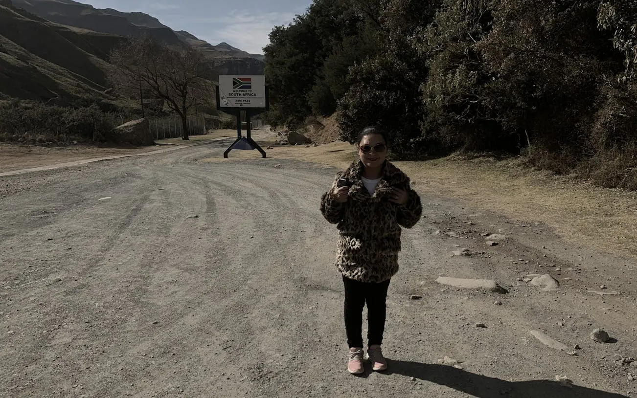 Person standing near Sani Pass sign on a dirt road, surrounded by mountains and trees.