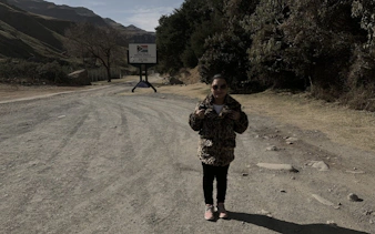 Person standing near Sani Pass sign on a dirt road, surrounded by mountains and trees.