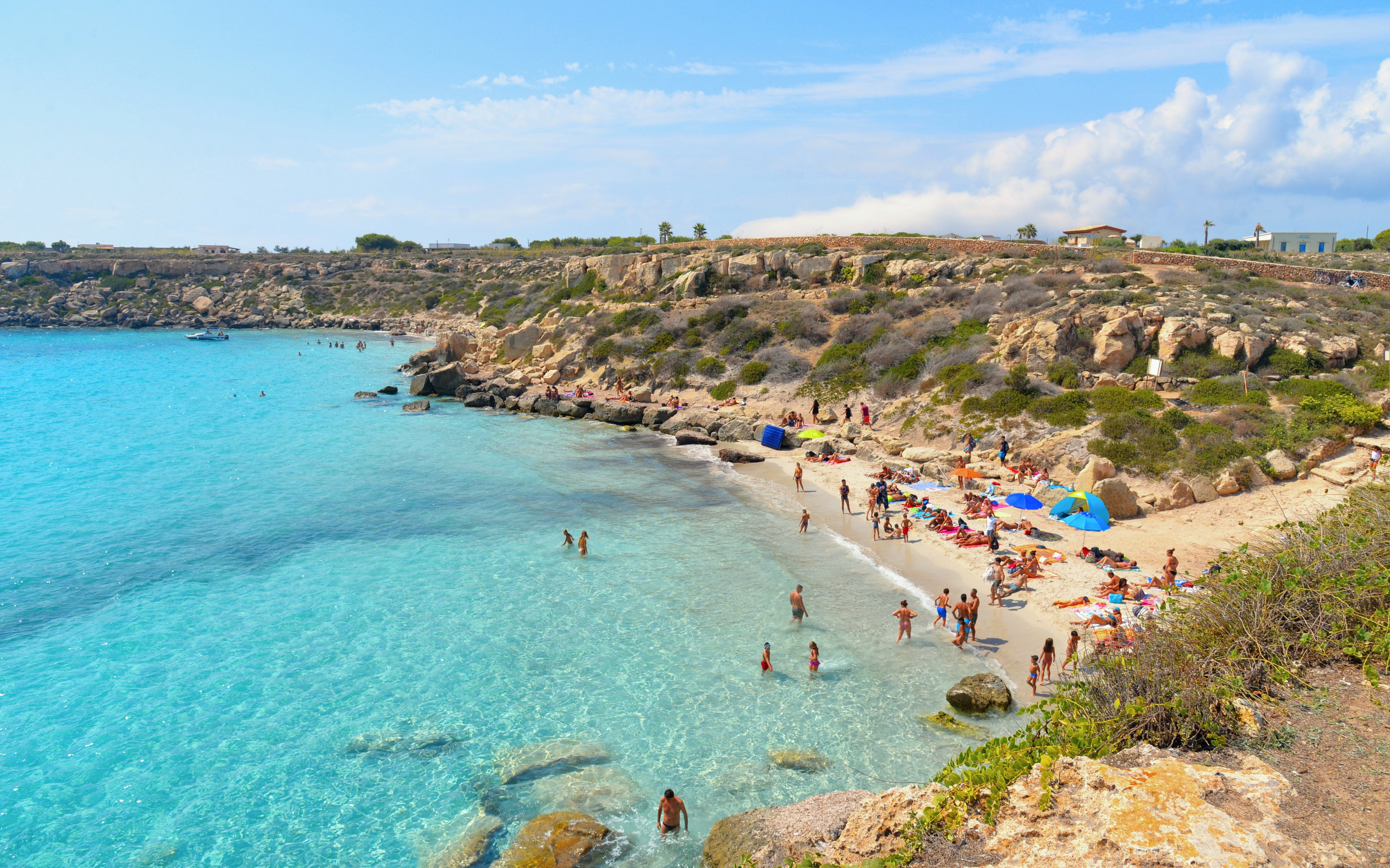 Turquoise blue water and rocky beach on Favignana Island with people swimming and sunbathing.
