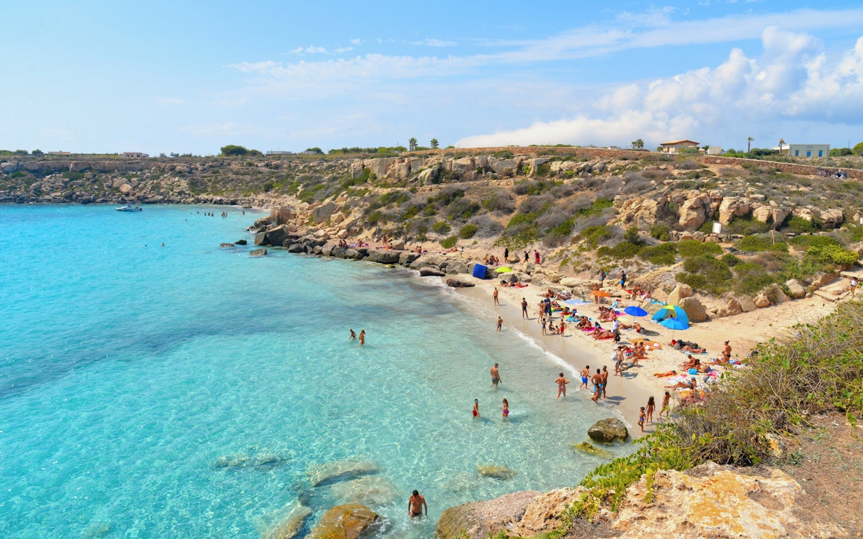 Turquoise blue water and rocky beach on Favignana Island with people swimming and sunbathing.