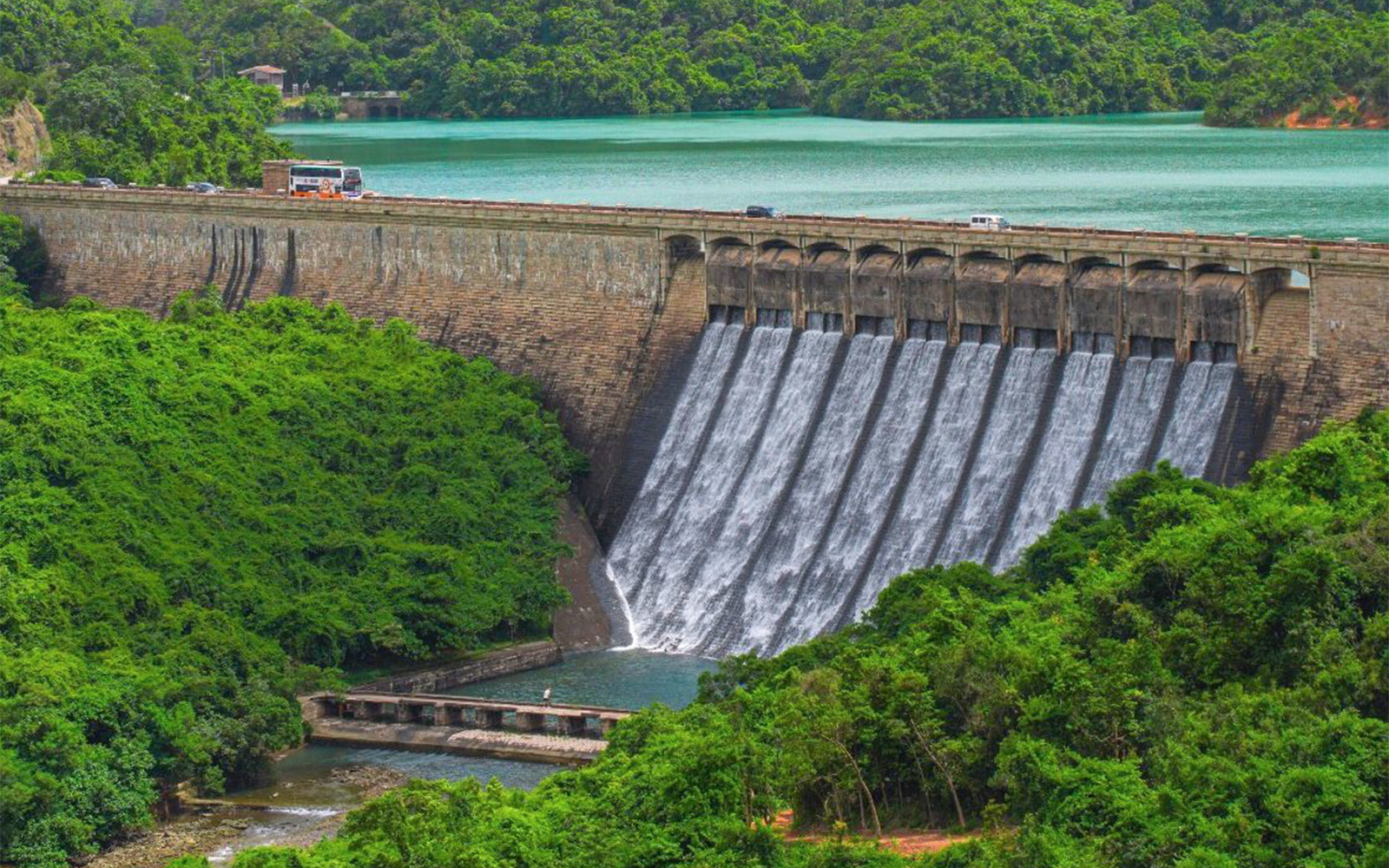 Big Bus tour passing over a dam with cascading water and lush greenery.