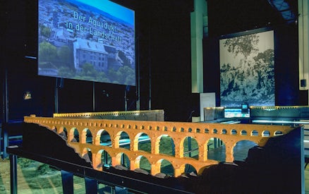 Model of Pont du Gard aqueduct in museum exhibit with cinema screen in background.