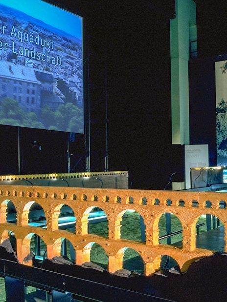 Model of Pont du Gard aqueduct in museum exhibit with cinema screen in background.