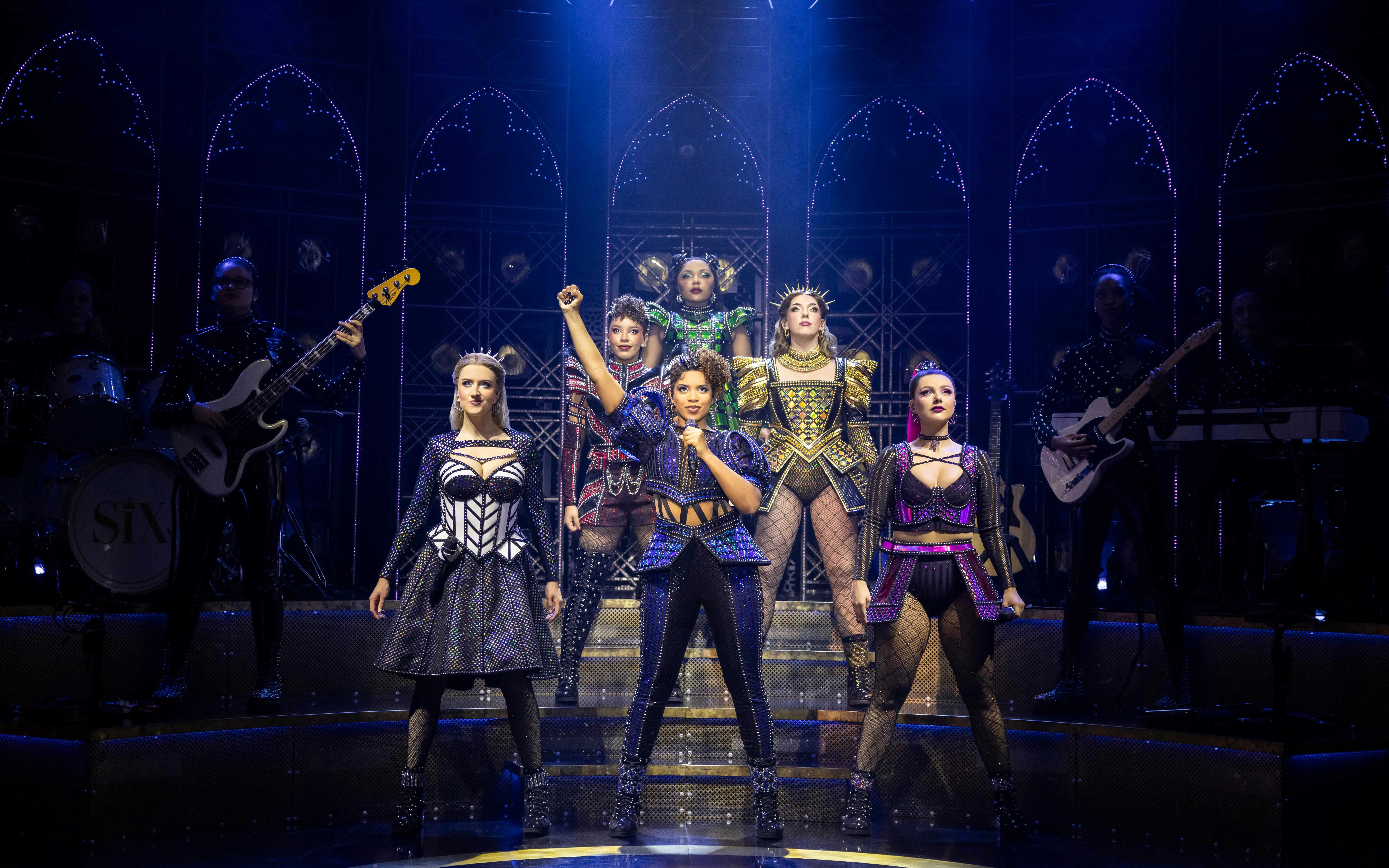 Cast of Six performing on West End stage with vibrant costumes and dynamic lighting.