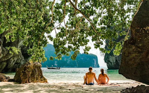 Couple sitting on Hong Island beach, Krabi, with speedboat and cliffs in view.