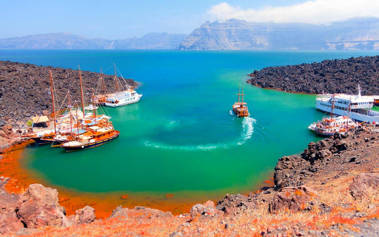Boats anchored near Nea Kameni volcanic island, Santorini, with rocky landscape and turquoise water.
