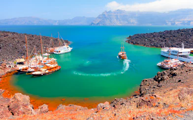 Boats anchored near Nea Kameni volcanic island, Santorini, with rocky landscape and turquoise water.