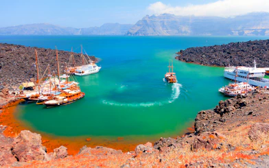 Boats anchored near Nea Kameni volcanic island, Santorini, with rocky landscape and turquoise water.