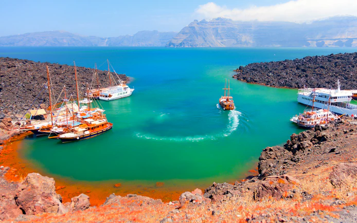 Boats anchored near Nea Kameni volcanic island, Santorini, with rocky landscape and turquoise water.