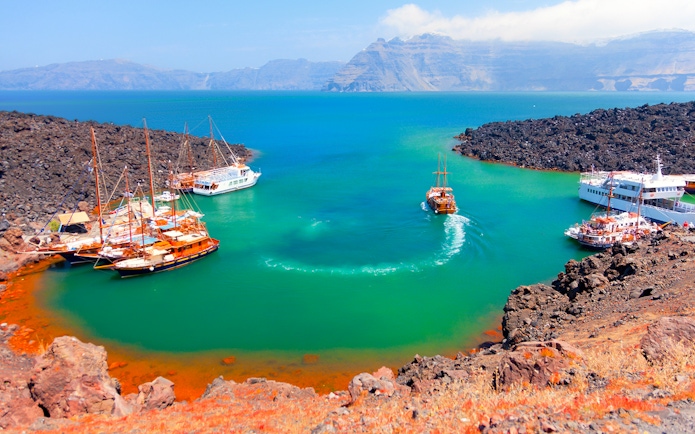 Boats anchored near Nea Kameni volcanic island, Santorini, with rocky landscape and turquoise water.