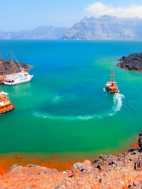 Boats anchored near Nea Kameni volcanic island, Santorini, with rocky landscape and turquoise water.