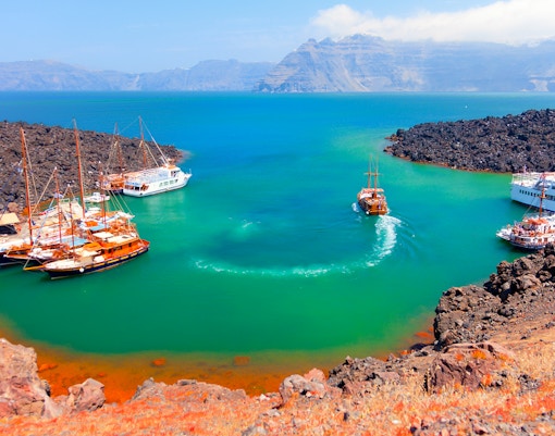 Boats anchored near Nea Kameni volcanic island, Santorini, with rocky landscape and turquoise water.
