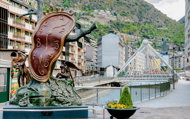Sculpture of melting clock in Andorra la Vella with bridge and mountains in background.