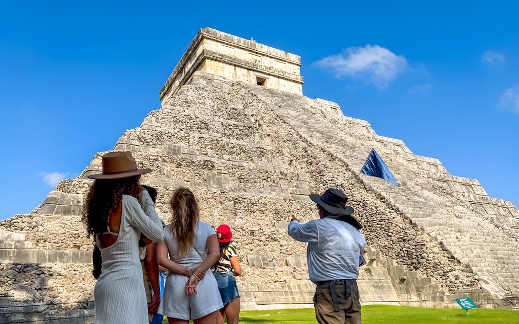 Tourist guide explaining Chichen Itza temple to visitors under a clear blue sky.