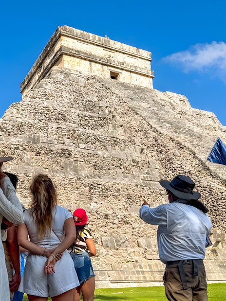 Tourist guide explaining Chichen Itza temple to visitors under a clear blue sky.