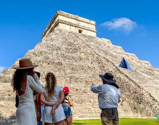 Tourist guide explaining Chichen Itza temple to visitors under a clear blue sky.