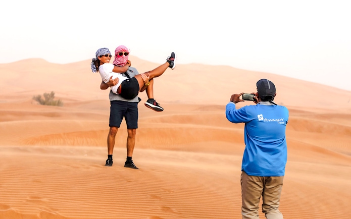 Couple posing in Abu Dhabi desert during safari, man lifting woman while guide takes photo.