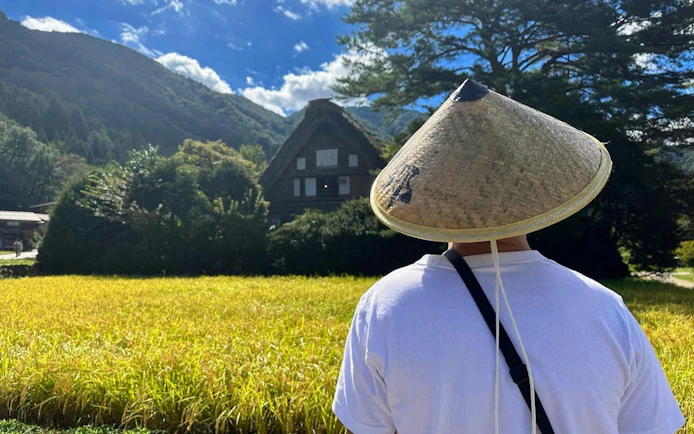 Person in traditional hat viewing Shirakawa-go farmhouse and rice field on Kanazawa day tour.