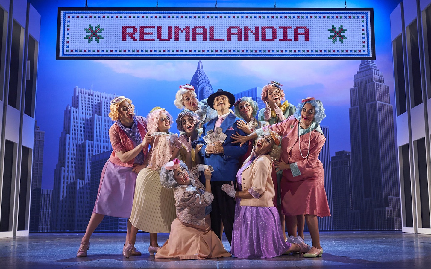 Cast of "The Producers" performing on stage with New York City backdrop.