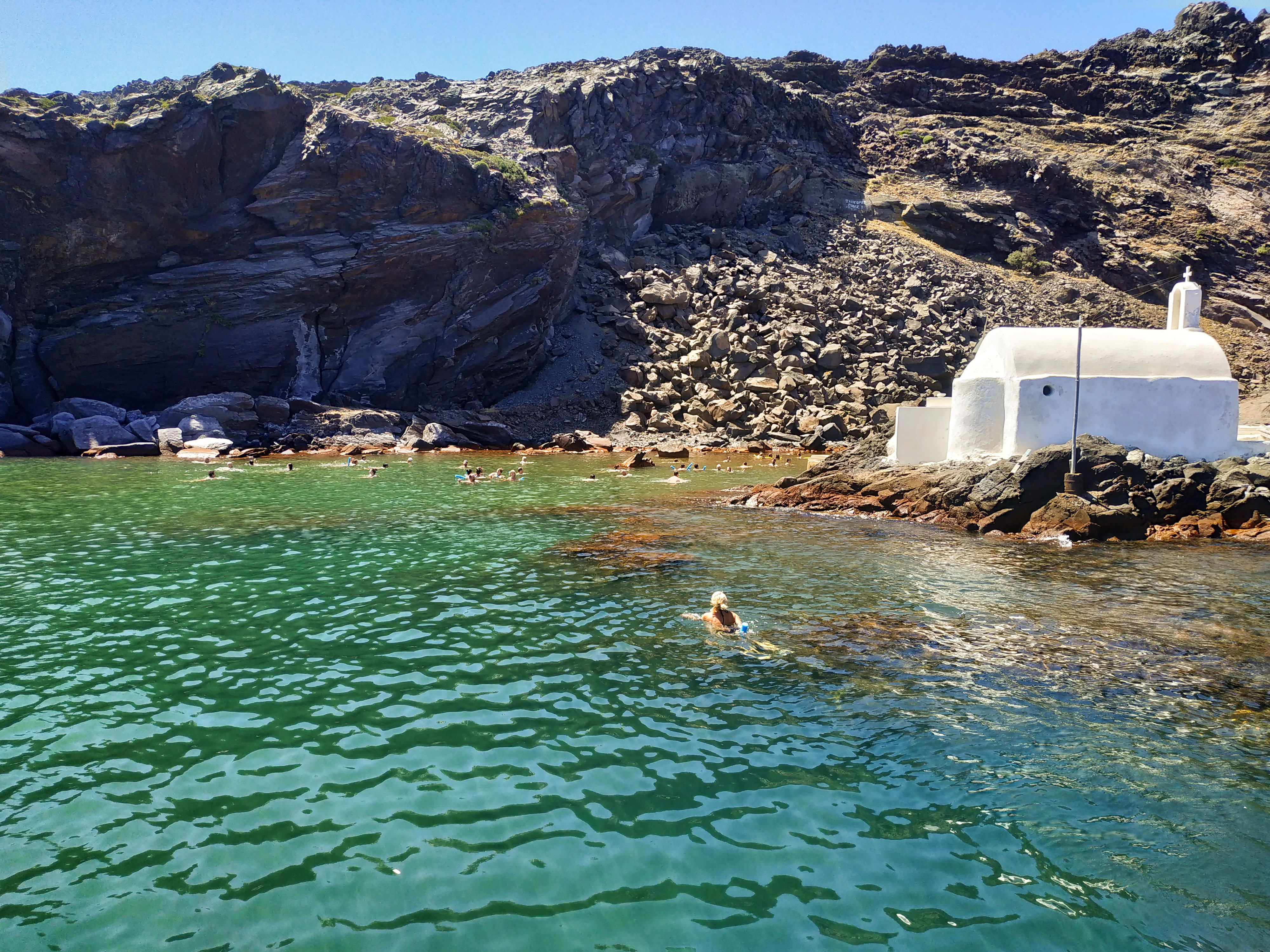 Swimmers near the rocky coast and white chapel of Nea Kameni, Santorini.