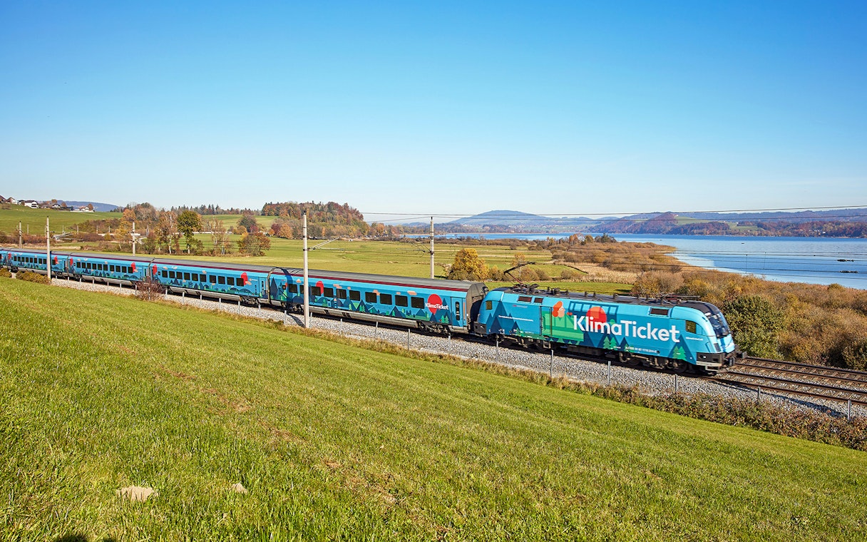 Train traveling through scenic countryside near a lake, promoting KlimaTicket.