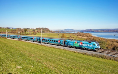 Train traveling through scenic countryside near a lake, promoting KlimaTicket.