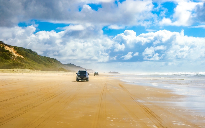 Four-wheel drive vehicles on Fraser Island beach, K'gari, with distant shipwreck.