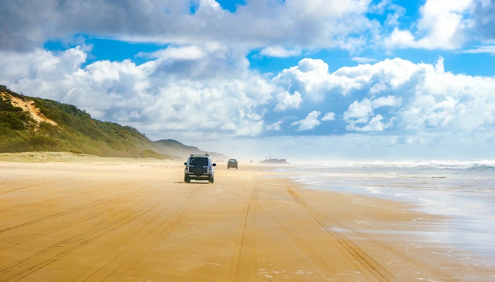 Four-wheel drive vehicles on Fraser Island beach, K'gari, with distant shipwreck.