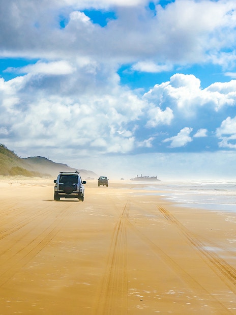 Four-wheel drive vehicles on Fraser Island beach, K'gari, with distant shipwreck.