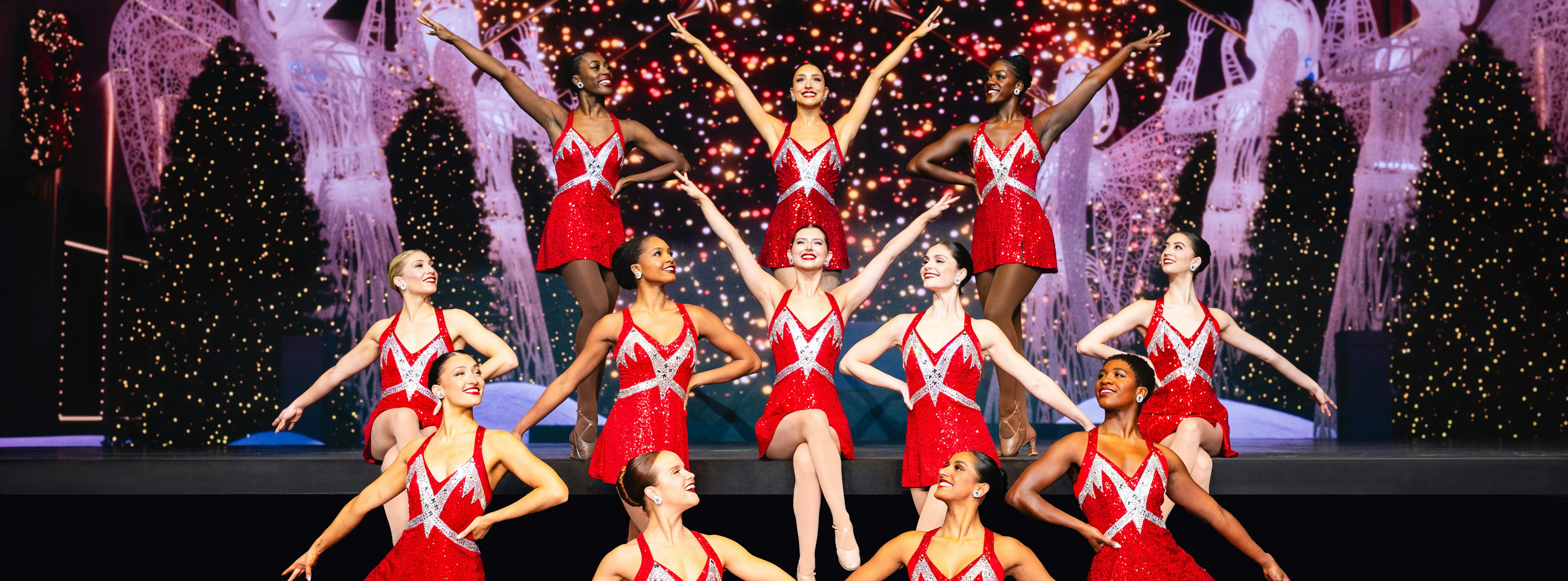 Rockettes performing in red costumes with Christmas lights and decorations in the background.