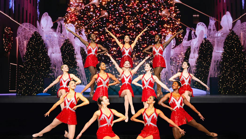 Rockettes performing in red costumes with Christmas lights and decorations in the background.