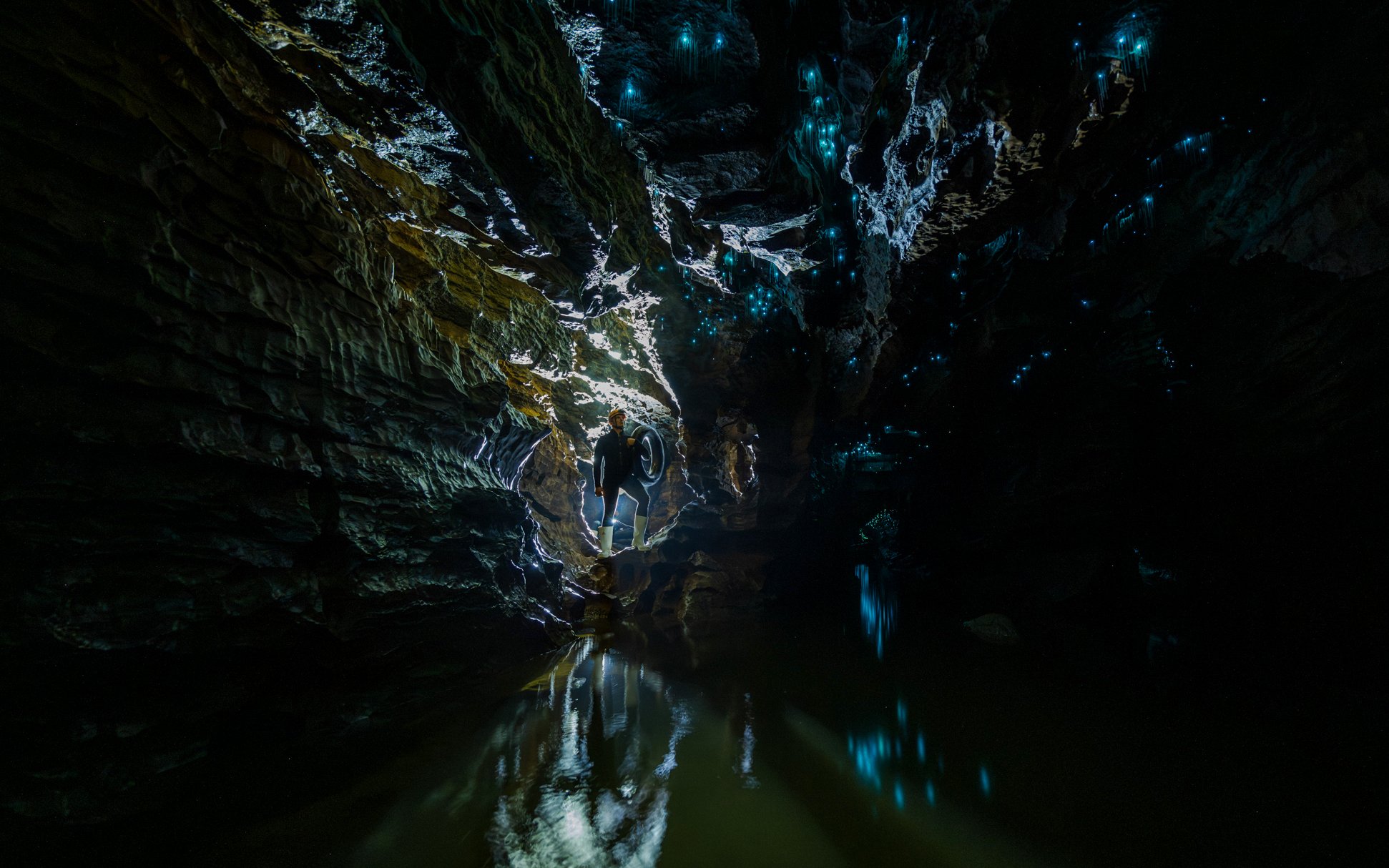 Person exploring Okohua Glowworm Cave with glowing blue lights on cave walls, Waitomo Experience.