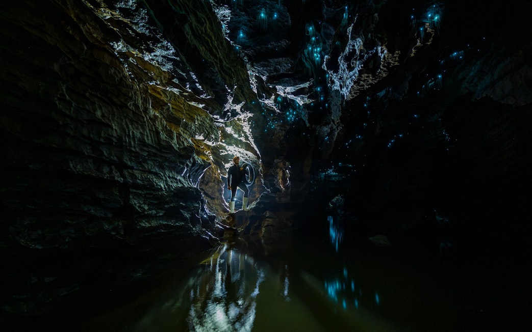 Person exploring Okohua Glowworm Cave with glowing blue lights on cave walls, Waitomo Experience.
