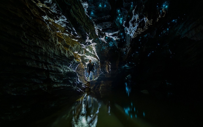 Person exploring Okohua Glowworm Cave with glowing blue lights on cave walls, Waitomo Experience.