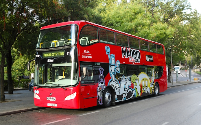 Red double-decker hop-on hop-off bus in Madrid near historic buildings.