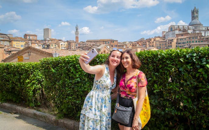 Tourists taking a selfie with Siena skyline in the background, featuring Torre del Mangia, Tuscany.