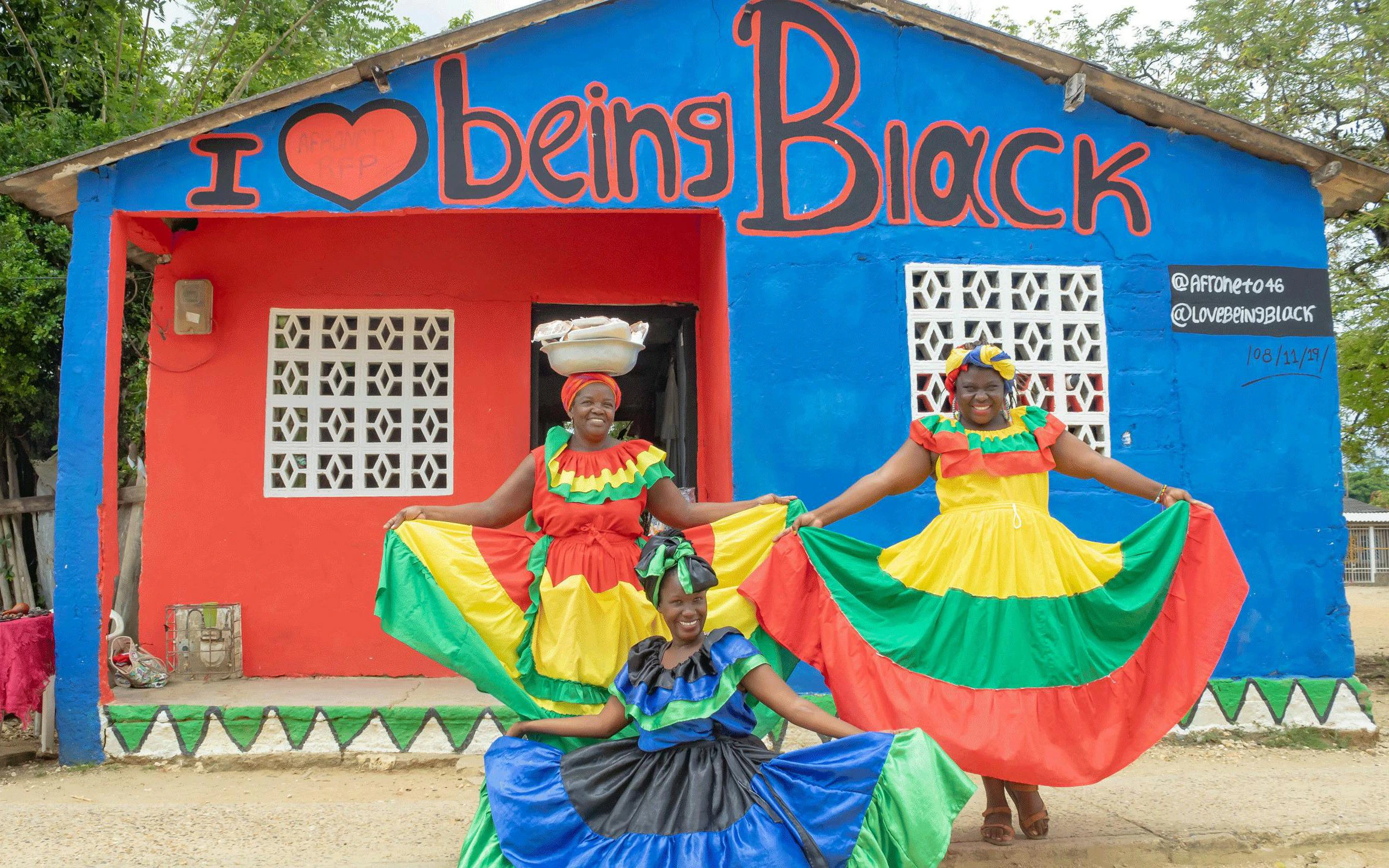 Traditional dancers in colorful dresses pose in front of San Basilio de Palenque mural.