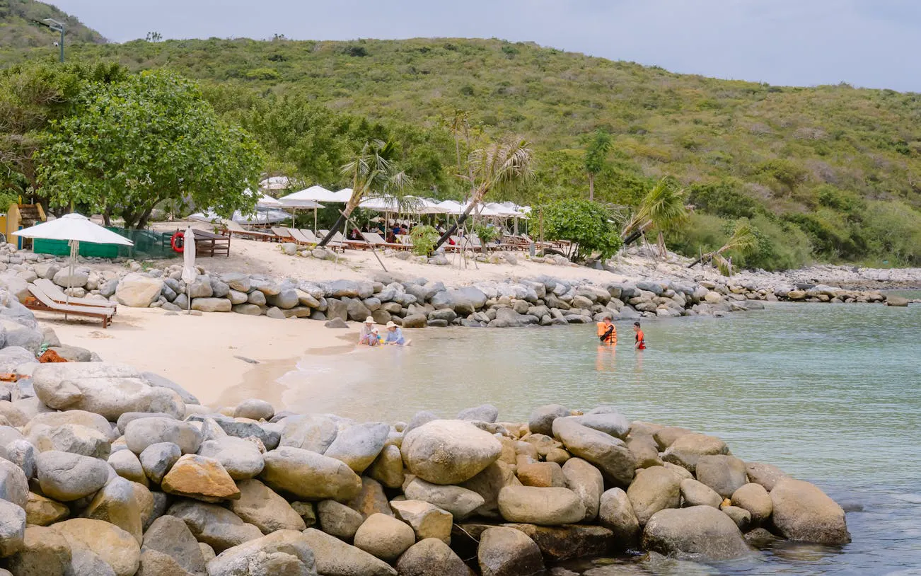 Nha Trang beach with sun loungers, trees, and people swimming near rocky shore.