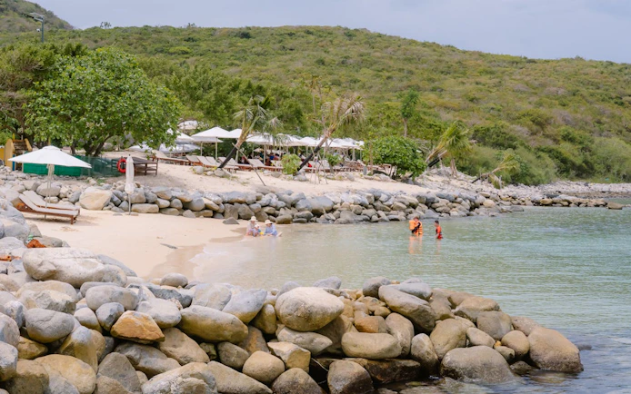 Nha Trang beach with sun loungers, trees, and people swimming near rocky shore.