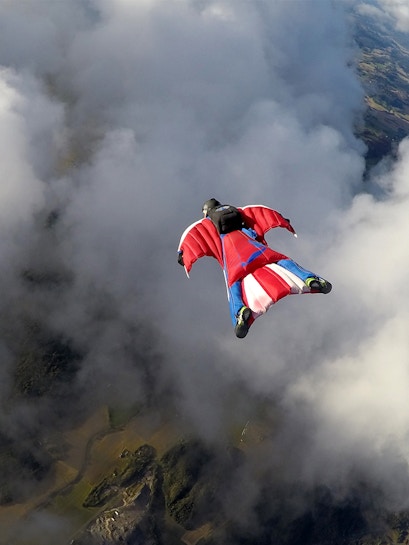 Skydiver in a wingsuit gliding through clouds over a scenic landscape.