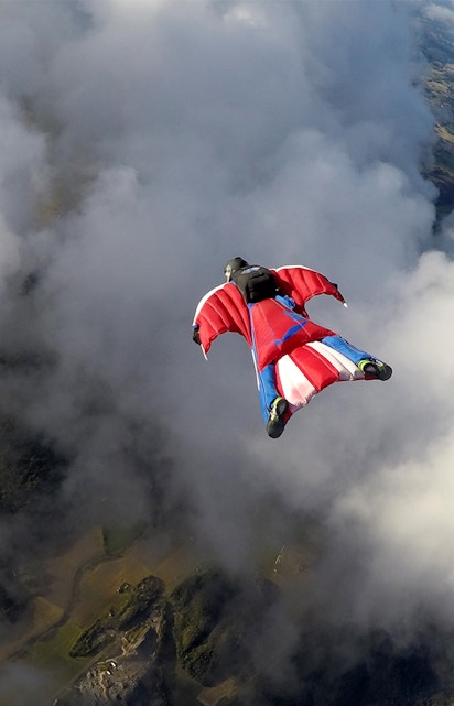 Skydiver in a wingsuit gliding through clouds over a scenic landscape.