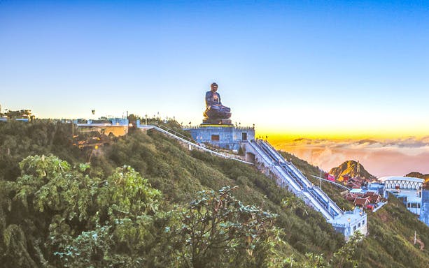 Great Amitabha Buddha statue on a hilltop at sunrise, surrounded by lush greenery.