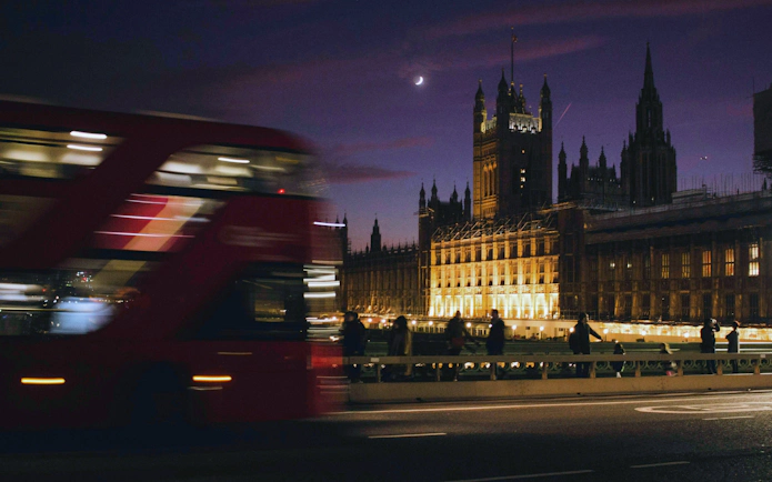 Red double-decker bus passing by the illuminated Houses of Parliament in London at night.