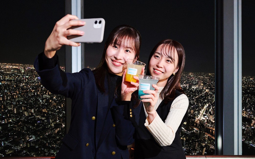Tourists taking a selfie with drinks on Tembo Deck, Tokyo at night.