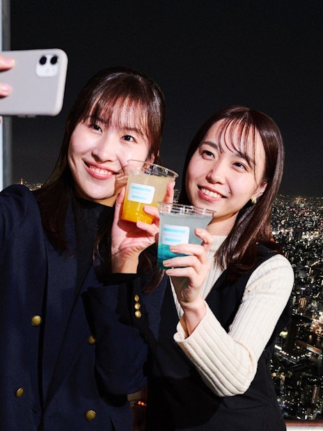 Tourists taking a selfie with drinks on Tembo Deck, Tokyo at night.